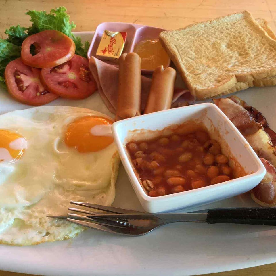A full English breakfast with fried eggs, bacon, sausages, baked beans, toast, and sliced tomatoes.