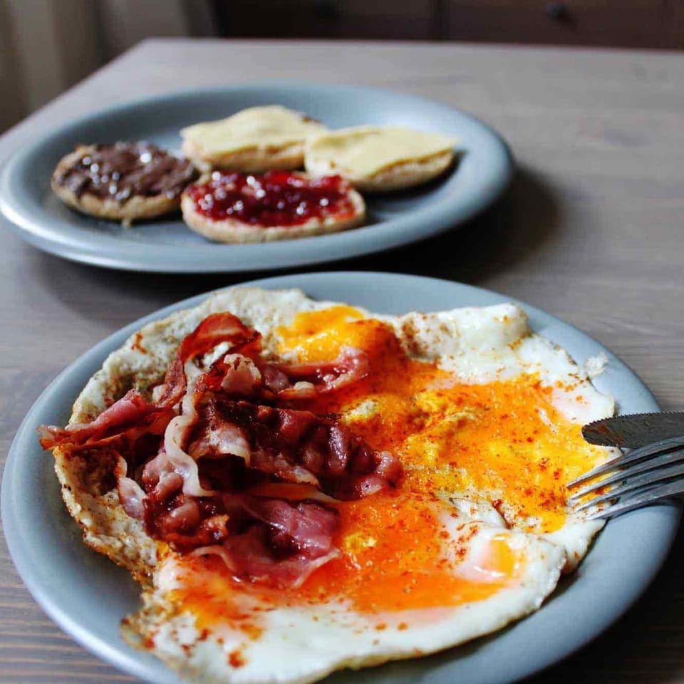A breakfast plate with fried eggs, bacon, and a separate plate of English muffins with spreads.