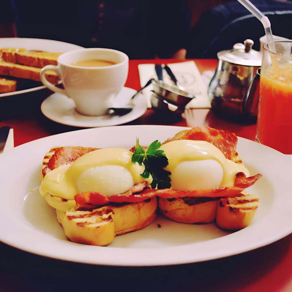Two eggs Benedict with bacon and hollandaise sauce, served with toast, coffee, and orange juice.